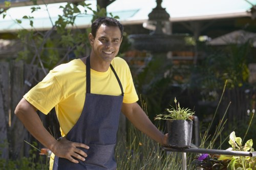 A man with dark hair and a warm smile stands in a well-maintained garden, wearing a bright yellow T-shirt and a dark apron. He is holding a small potted plant with green foliage, positioned on a garden table or surface. Behind him, the garden features neatly trimmed hedges, lush green grass, and a variety of garden plants, including tall grasses and flowering shrubs. In the background, there is a wooden trellis or fence, with greenery and flowering plants climbing or growing around it. The setting appears to be outdoors on a sunny day, with natural light illuminating the garden space, which exhibits a typical landscaped backyard or front garden layout with a mix of lawn, planting beds, and decorative elements suitable for garden maintenance or landscaping services in the Blackheath area.